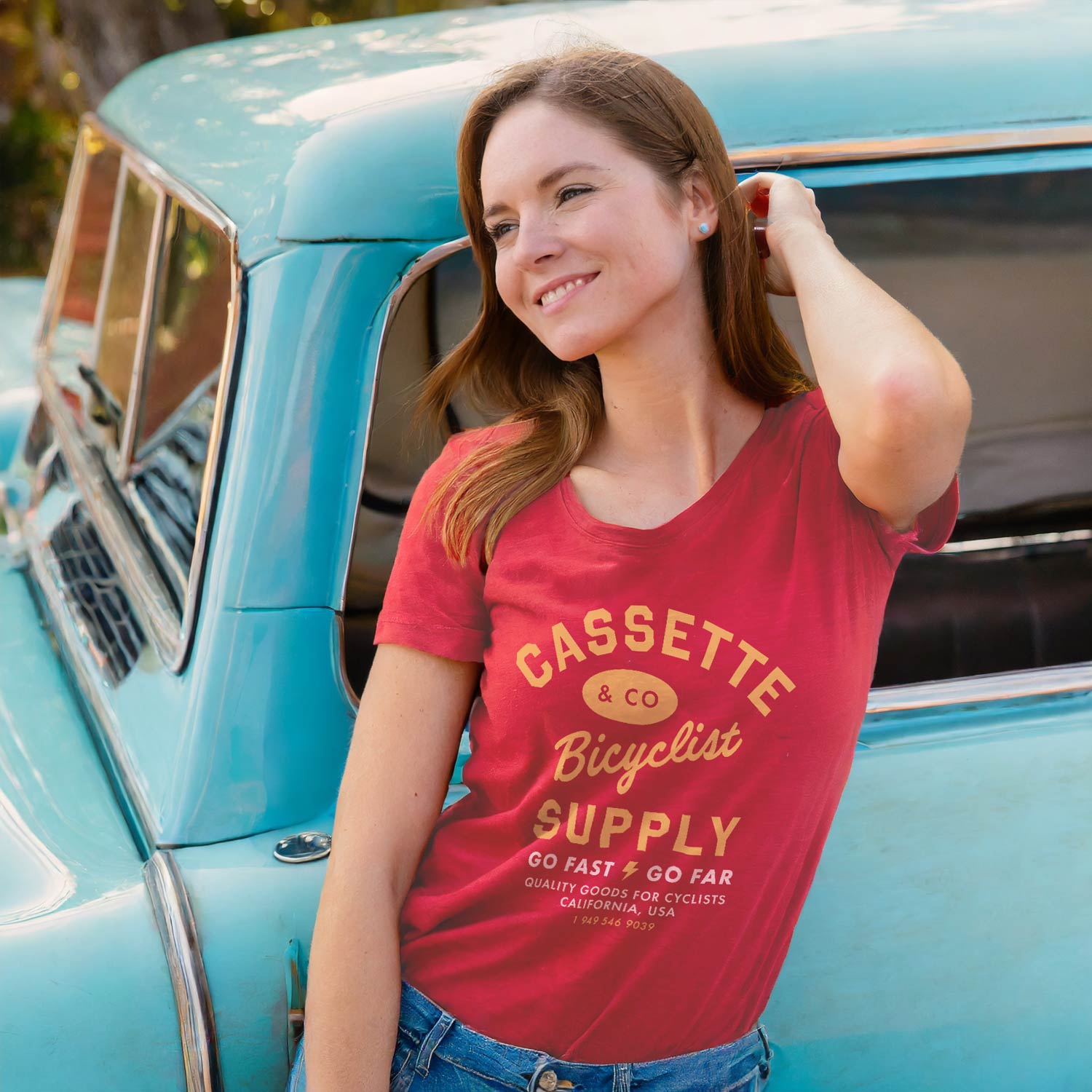 woman leans up against light blue vintage truck while wearing the go fast go far red women's tee